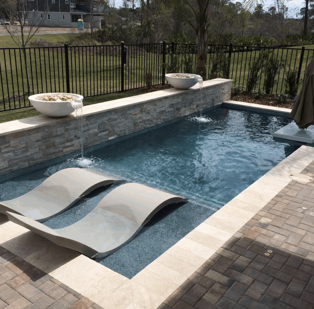 Rectangular backyard pool with two built-in lounge chairs, a tiled wall with two water bowls, and a stone paver patio, surrounded by a black metal fence.